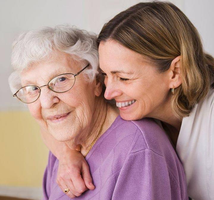 Caregiver Smiling resident looking at the camera while a caregiver hugs her from behind.