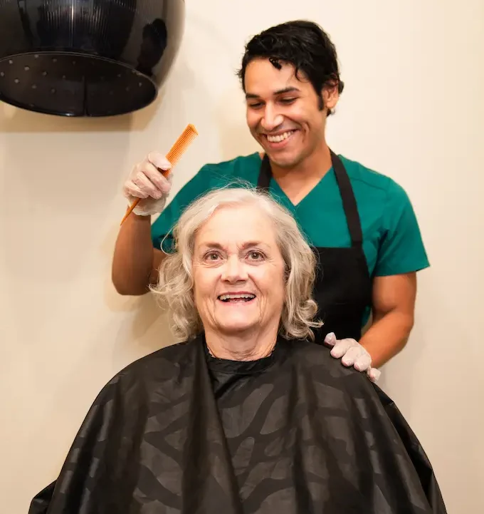 A Dementia care patient smiling while their hair is styled by a friendly hairdresser in an Avalon Memory Care uniform.
