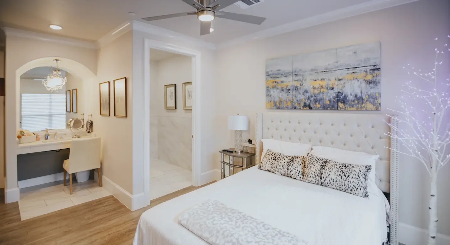 A spacious bedroom featuring a queen sized bed, a bathroom vanity with modern finishing, white decor, and contemporary artwork.
